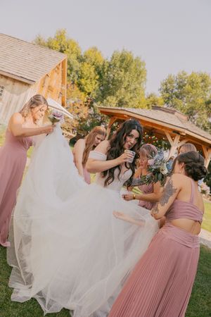 Bridesmaids in dusty rose dresses help fluff a white wedding gown outdoors.