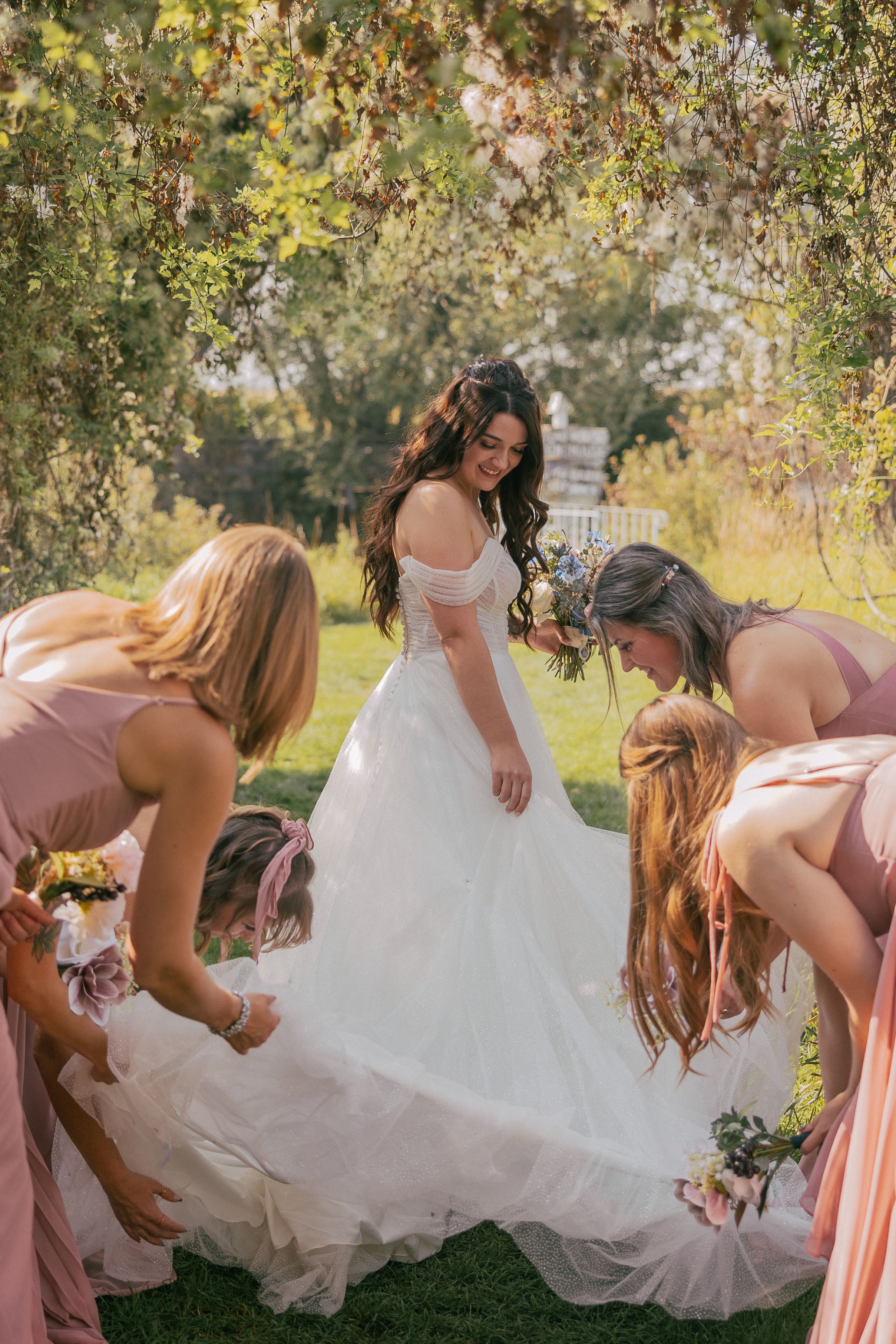 Wedding party in pink dresses playfully poses while looking under the bride's dress.