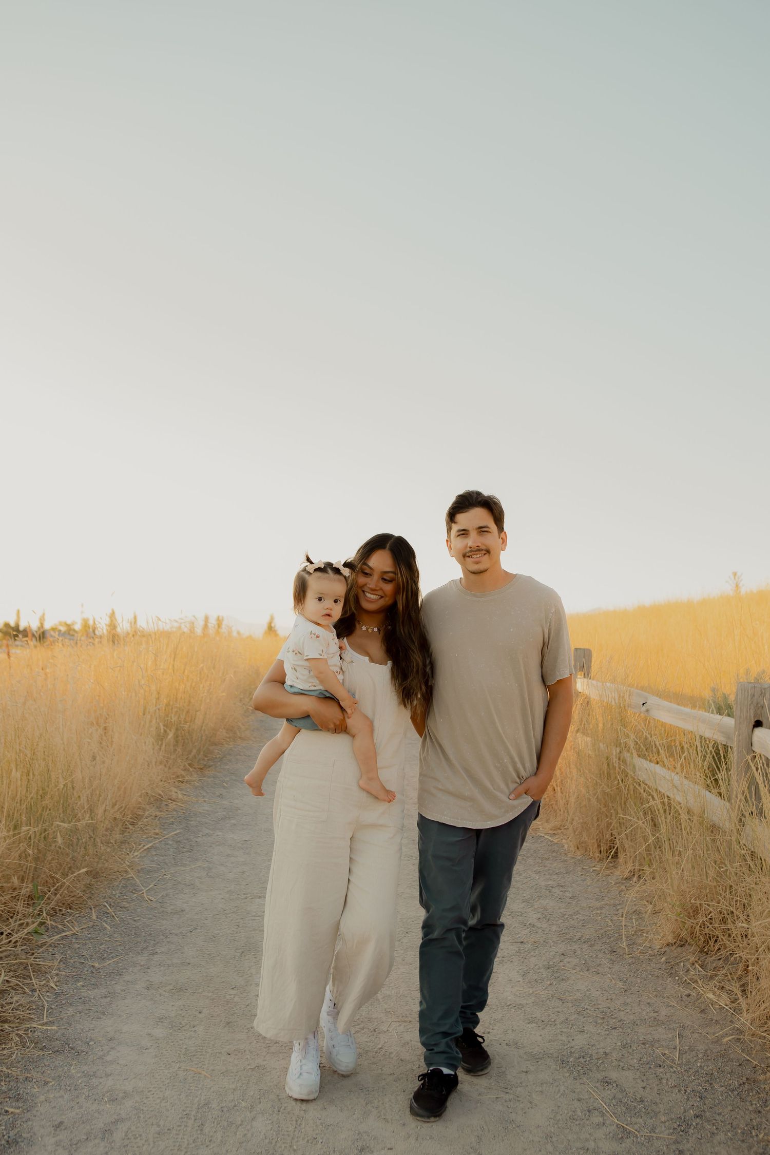 A family walks together at sunset on a dirt path bordered by golden grass and wooden fencing.
