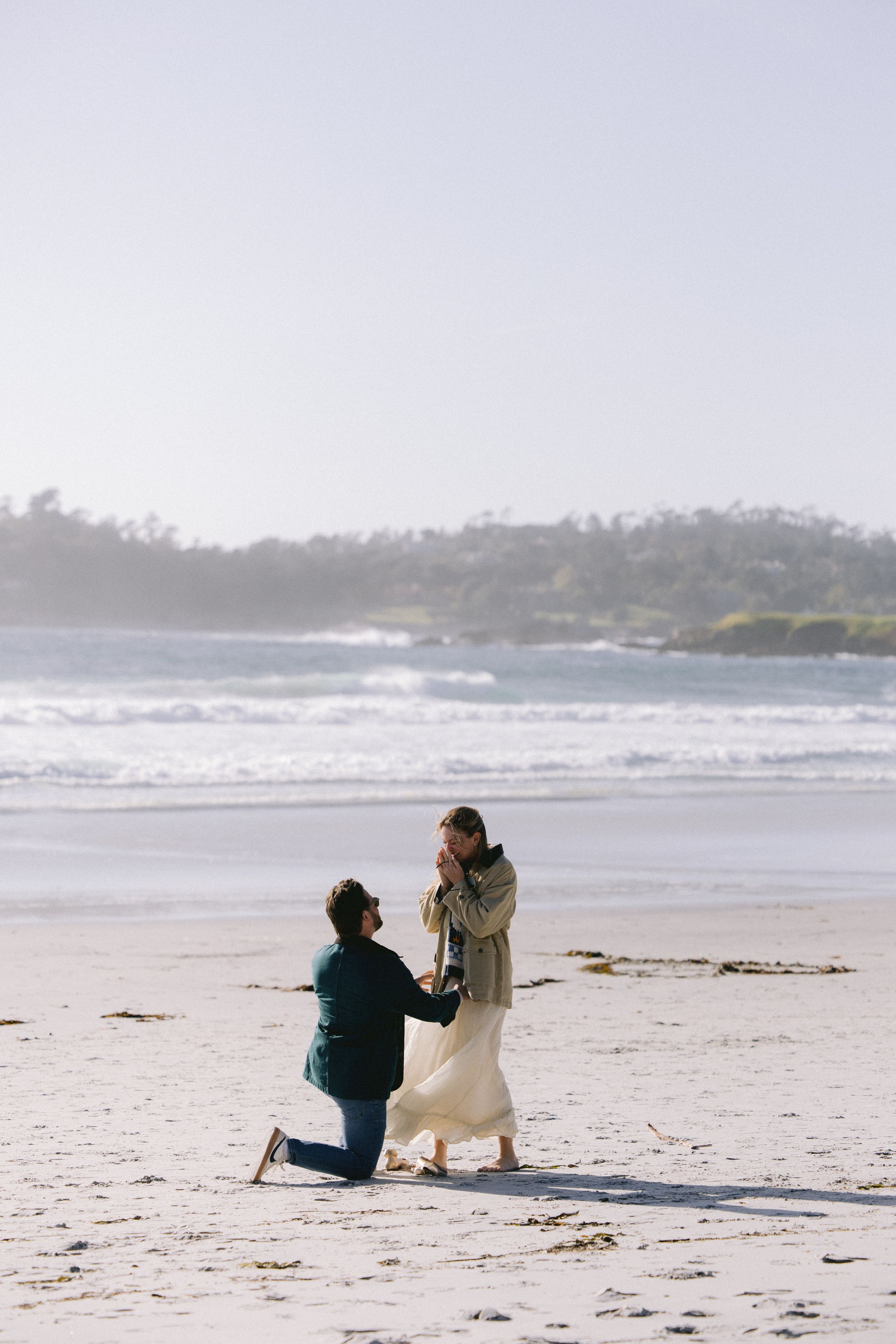 Two people sharing a romantic moment on a beautiful sandy beach with turquoise waves and green cliffs in background.