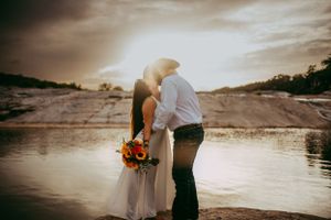 Elopement couple kissing at sunset with the rock formations and river behind them as she holds bouquet of sunflowers in Hill Country, Texas.