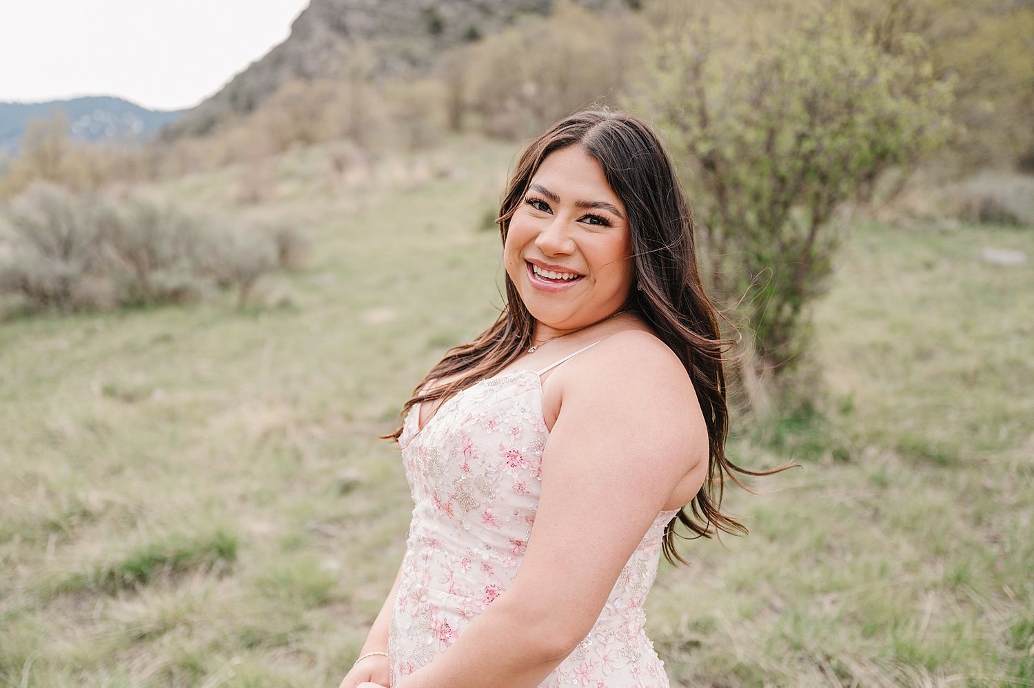 A person wearing a light pink formal dress smiles while posing in a grassy outdoor setting with mountains in the background.