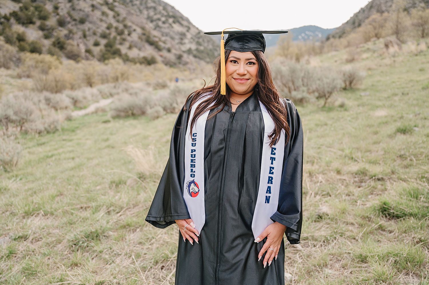 Graduate in cap and gown poses outdoors against mountainous desert landscape wearing graduation stole.