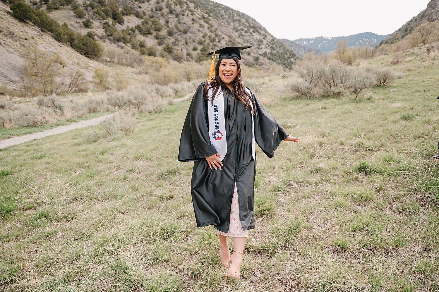 Graduate in cap and gown poses joyfully in a scenic mountain valley with desert landscape.