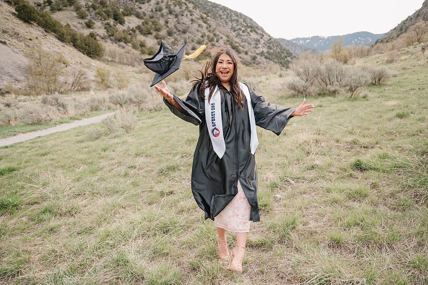Graduate celebrating in black graduation gown and stole raises cap joyfully in mountain valley setting.