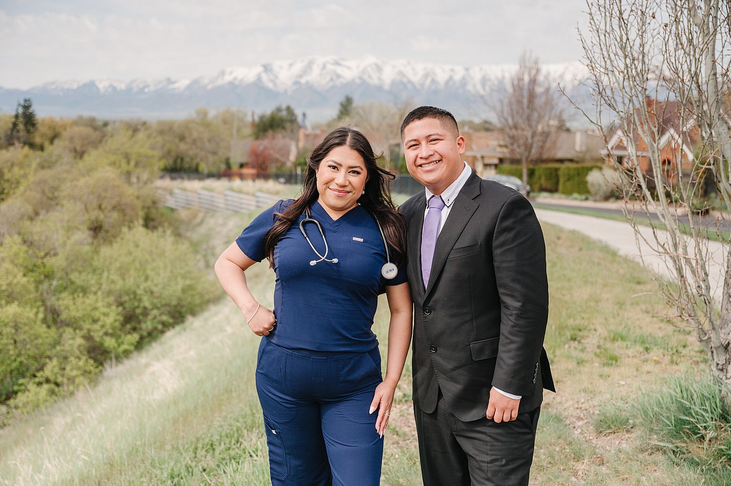 Healthcare professionals in navy blue scrubs and business attire stand together on a hilltop path with mountains in background.