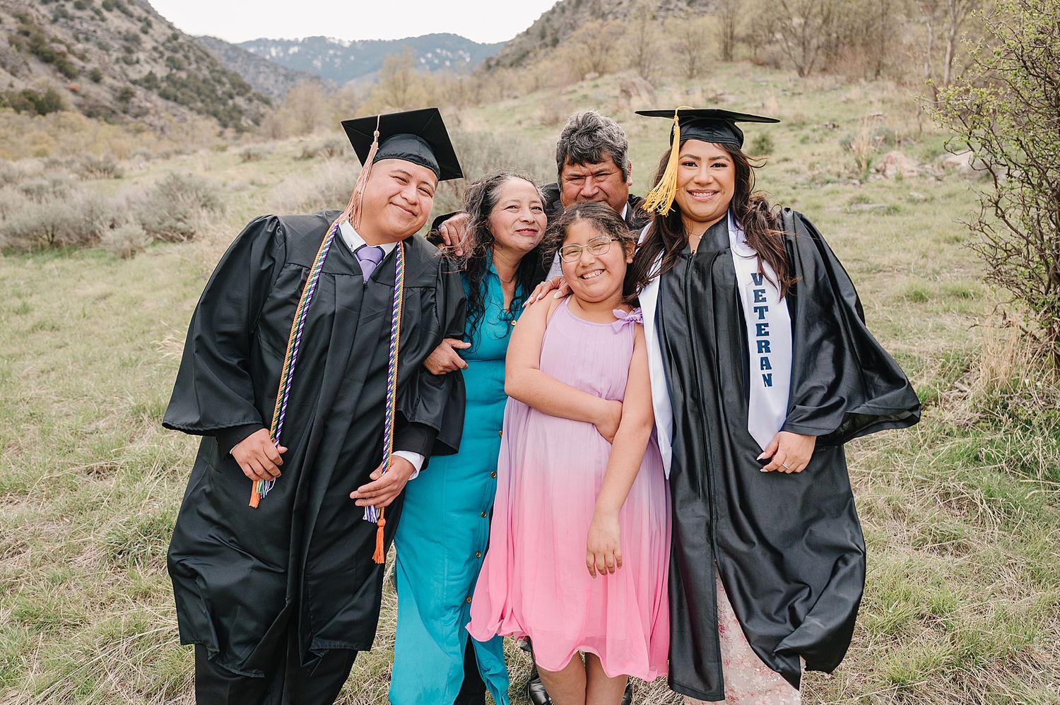 Group wearing graduation caps and gowns pose together on a hillside with mountains in the background.
