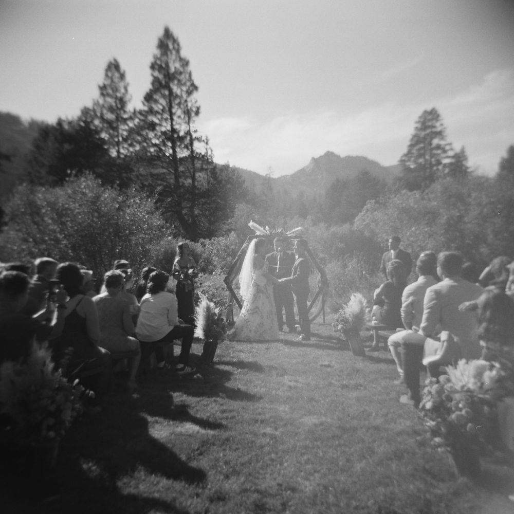 Black and white photo of outdoor wedding ceremony with mountain backdrop and gathered guests.