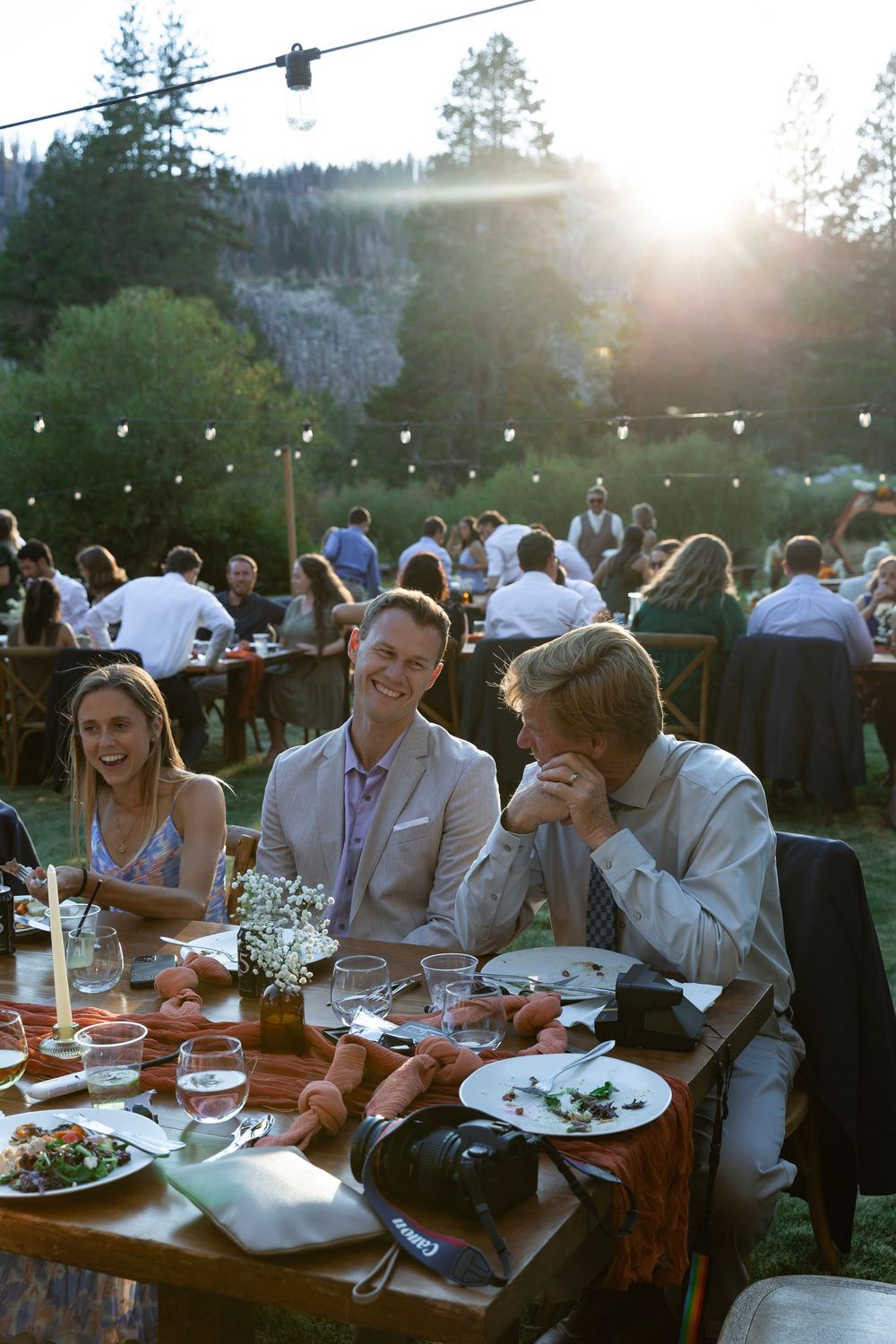 Wedding guests enjoying dinner at outdoor tables with string lights overhead at sunset.