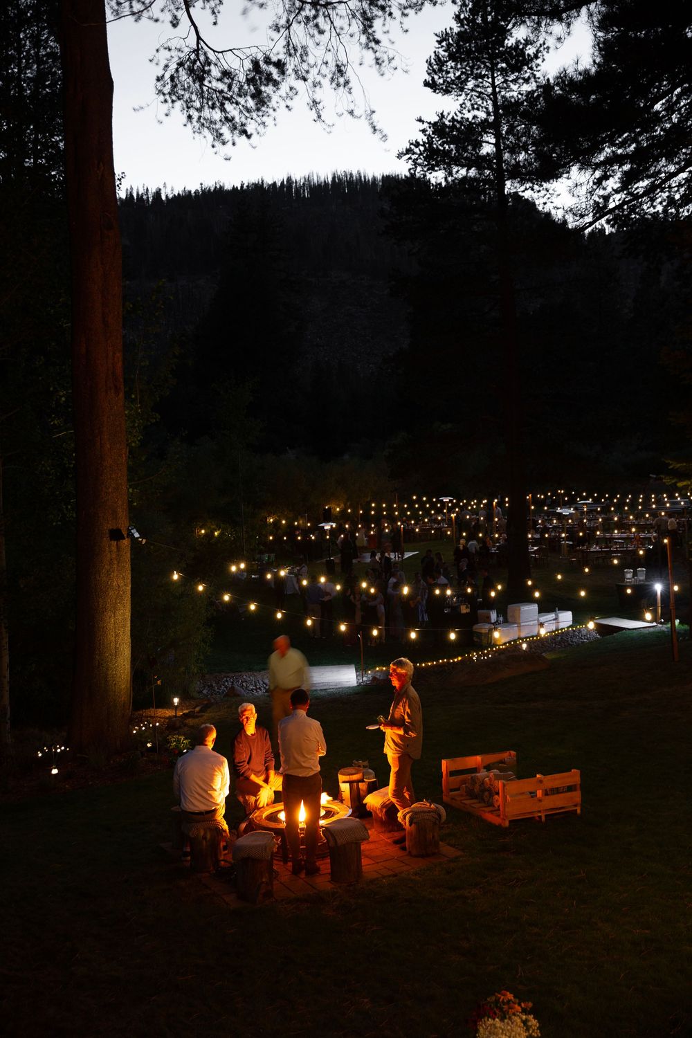 Group gathered around campfire with string lights decorating outdoor evening event.