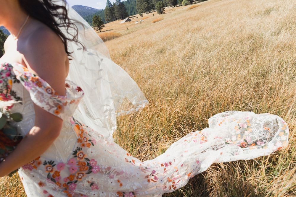 A floral wedding dress billows in the wind against a backdrop of sun-dried grass and mountains.