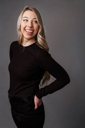 Professional studio portrait with black sweater and jeans against gray background showing joyful expression and dynamic pose.