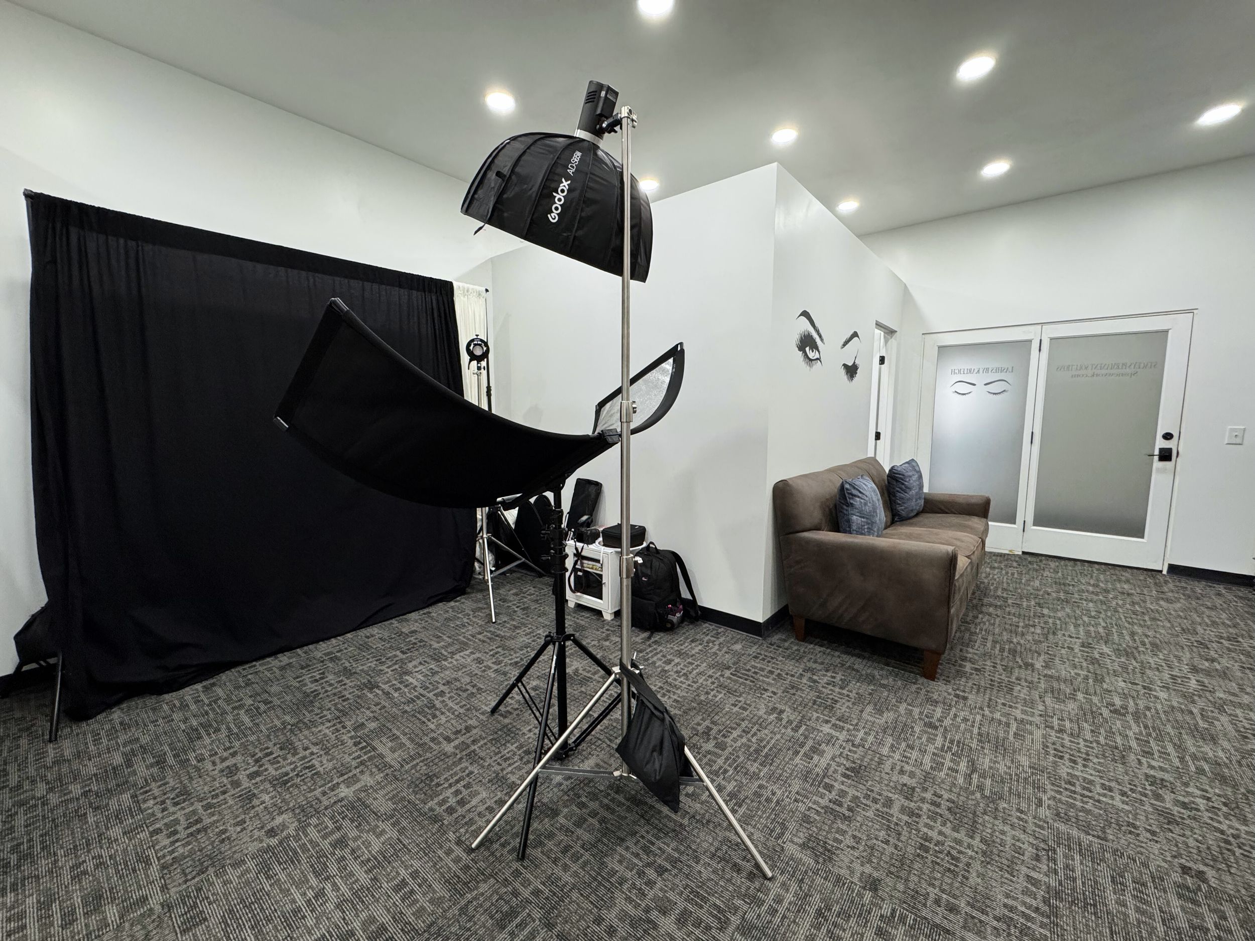 Modern photo studio interior with white walls, gray carpeted floor, lighting equipment and black backdrop setup.
