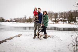 A group stands in the snow with their dog by a frozen lake on a winter day while houses dot the background.
