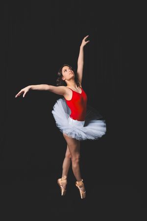 Ballerina in red leotard and white tutu performs graceful ballet pose on pointe shoes against dark background.