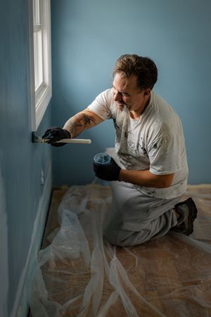 A person in white clothing crouches on a wooden floor while painting a blue wall in a room with natural light.