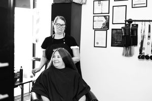 Black and white photo of a hairstylist working with client in salon with framed certificates on wall.