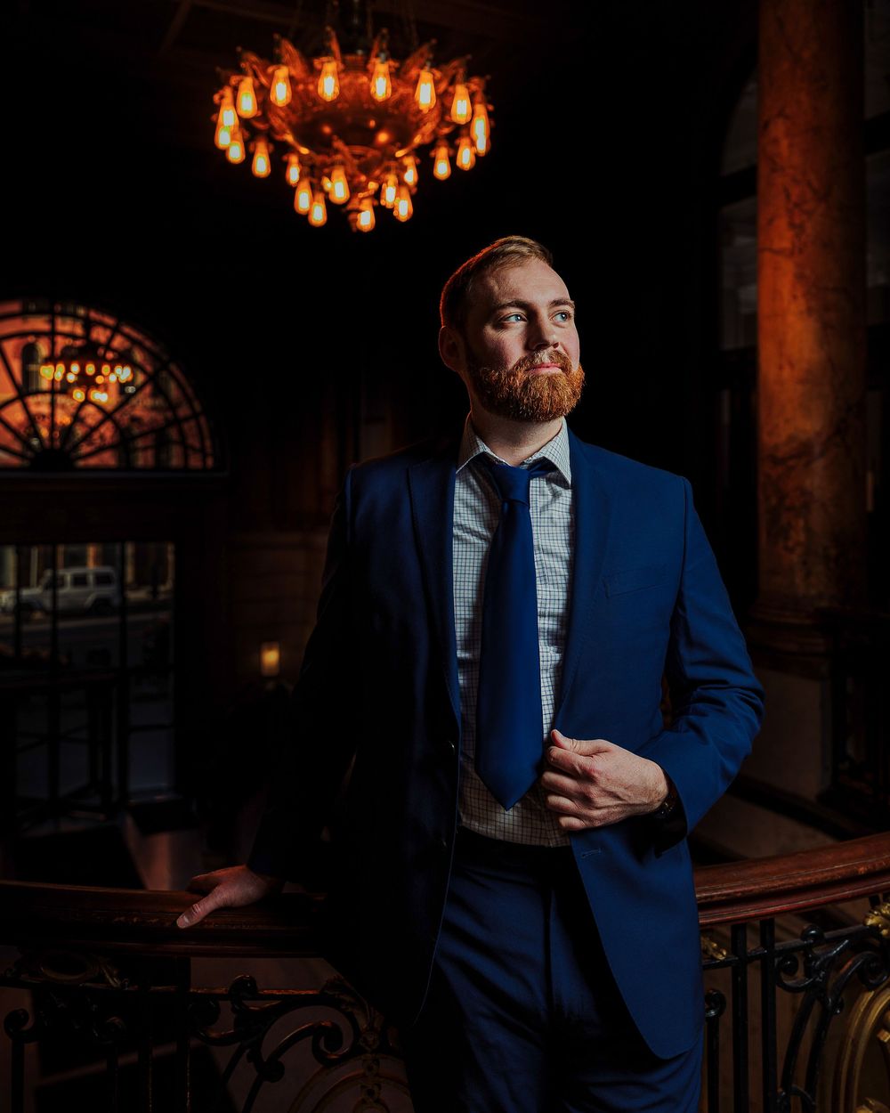 A person in a navy blue suit and tie poses in a dimly lit elegant room with an ornate chandelier overhead.