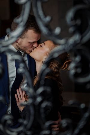 Romantic couple sharing an intimate kiss viewed through decorative wrought iron scrollwork in moody lighting.