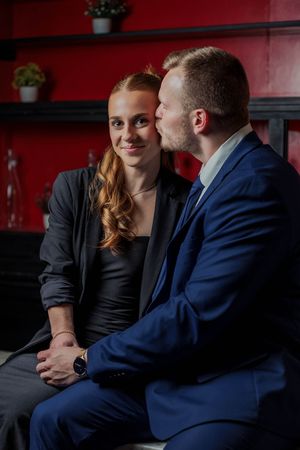 A romantic couple sharing an intimate moment on a bench against a red wall with potted plants in the background.