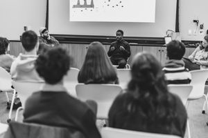 Black and white photo of students listening to a speaker in a classroom with a projection screen.