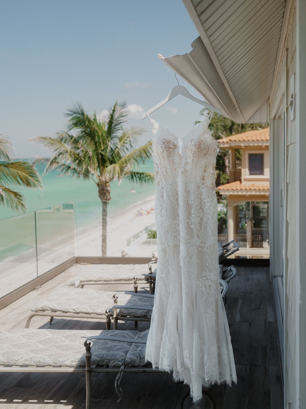 White lace wedding dress hanging on balcony with ocean and palm trees in background.