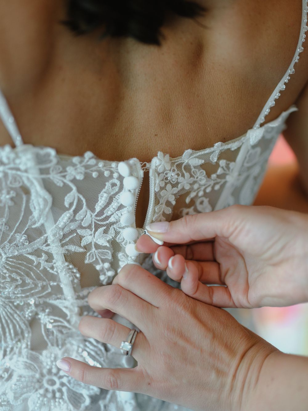 Hands carefully fastening small pearl buttons along the back of an intricate lace wedding gown.