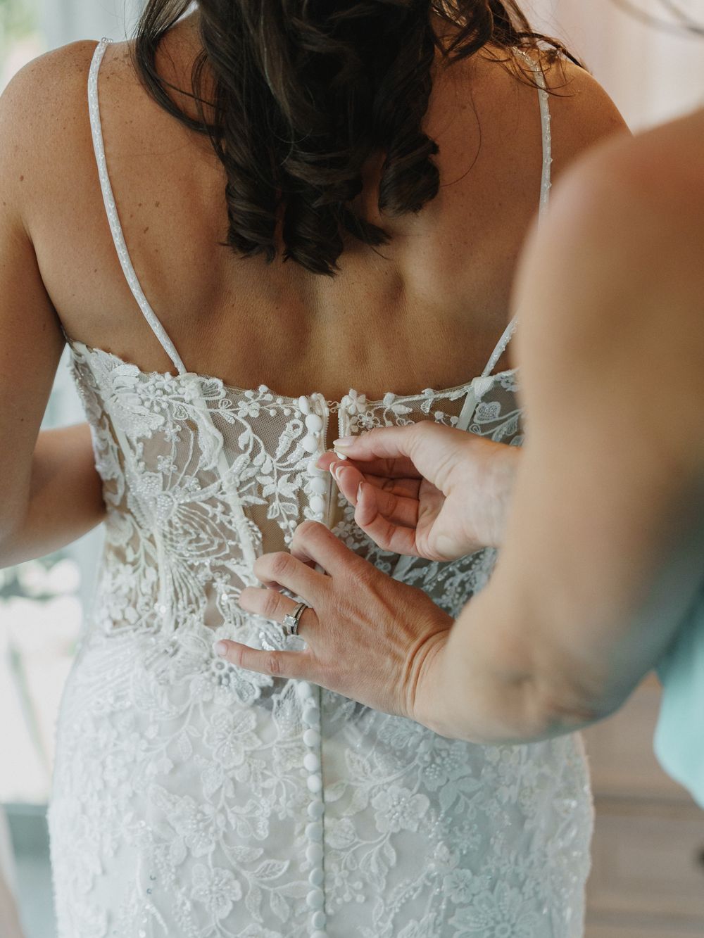 Close-up detail shots of delicate lace and buttons being fastened on a white wedding dress.