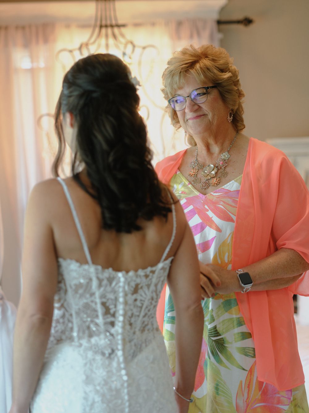 Helper in coral cardigan assisting with the wedding dress preparation in a sunlit room.