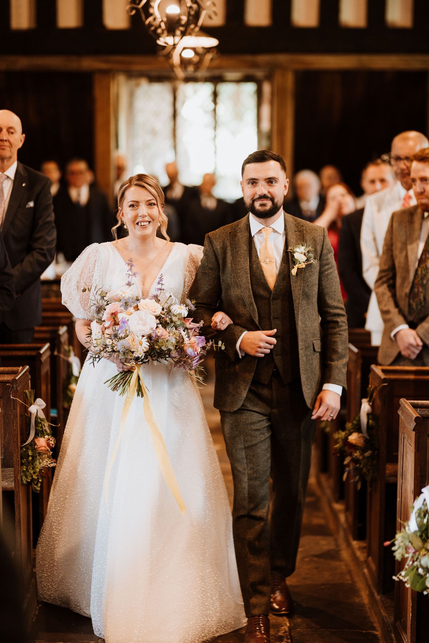 Bride and brother walks down a church aisle during their wedding ceremony, with guests watching from wooden pews.