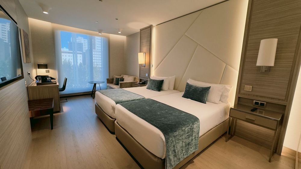 Modern hotel twin bedroom with beige decor, blue accent pillows, and large window.