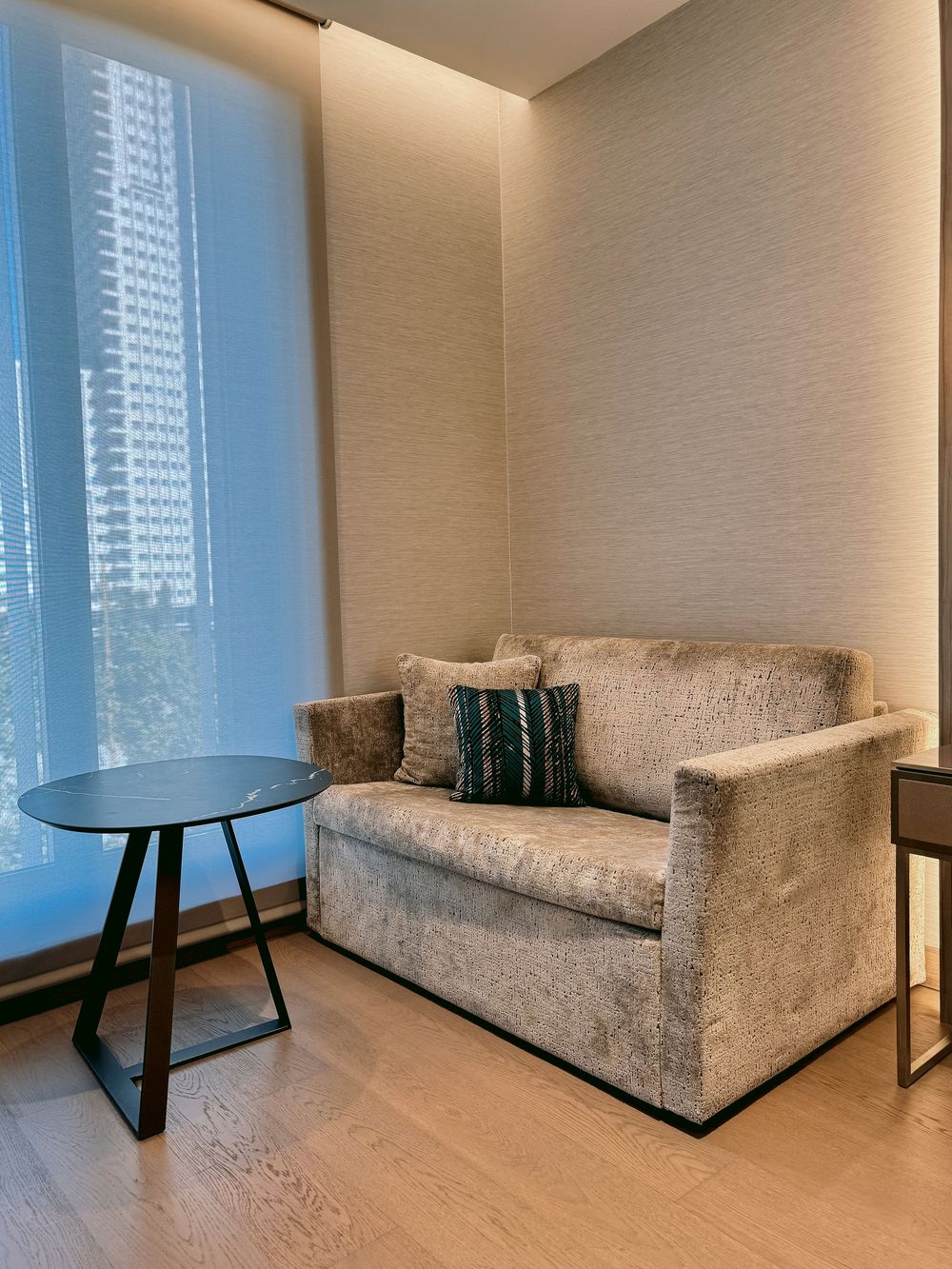 Elegant hotel room sitting area with grey loveseat sofa, side table, and floor-to-ceiling window.
