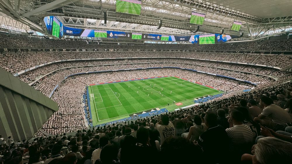 Packed football stadium with green pitch and large screens during a live match.
