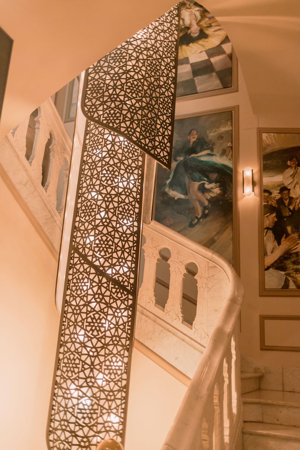 Ornate spiral staircase with decorative lattice railing and classical paintings on warm walls.
