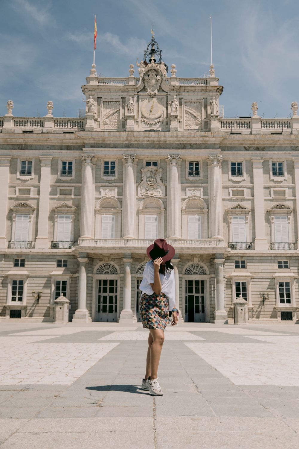 Woman in floral skirt and hat posing before the grand Royal Palace of Madrid on a sunny day.