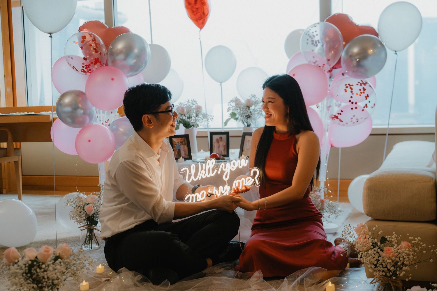 A romantic proposal setup with pink and blue balloons and a neon sign in a cozy indoor setting.