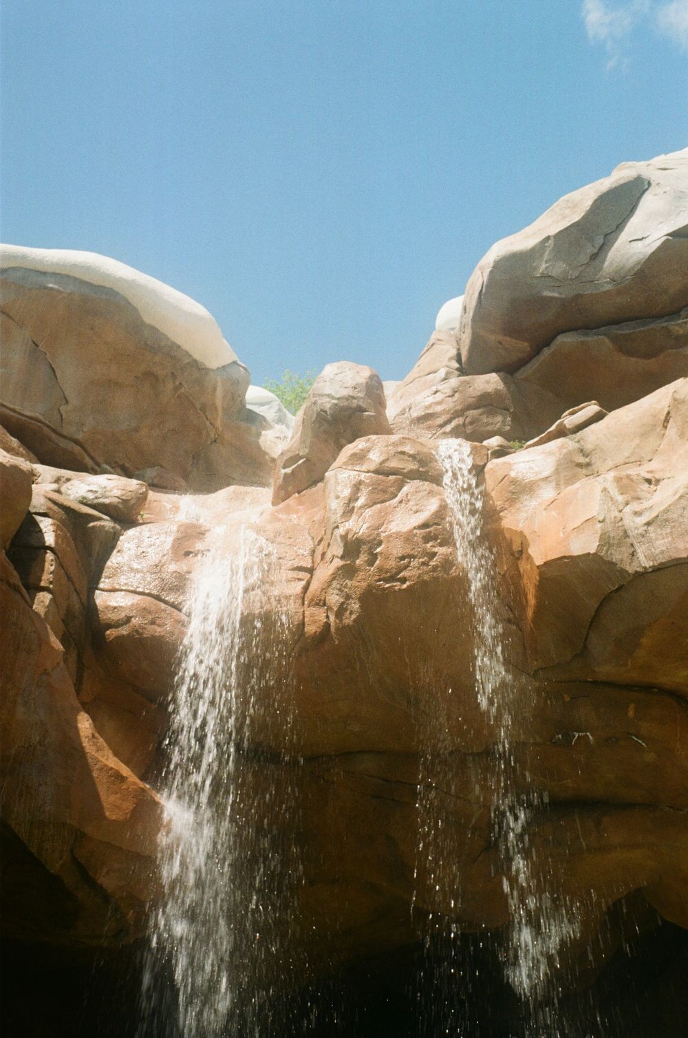 A waterfall cascades down tan sandstone rock formations against a bright blue sky in a desert landscape.