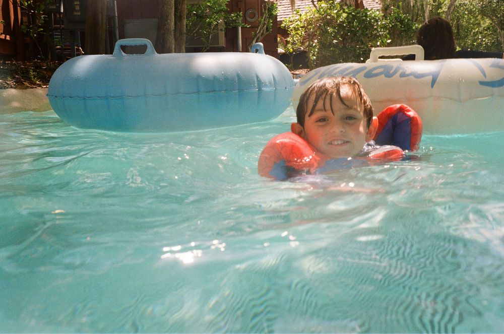 Small swimmer wearing orange water wings enjoys a refreshing dip in a bright turquoise pool on a sunny day.
