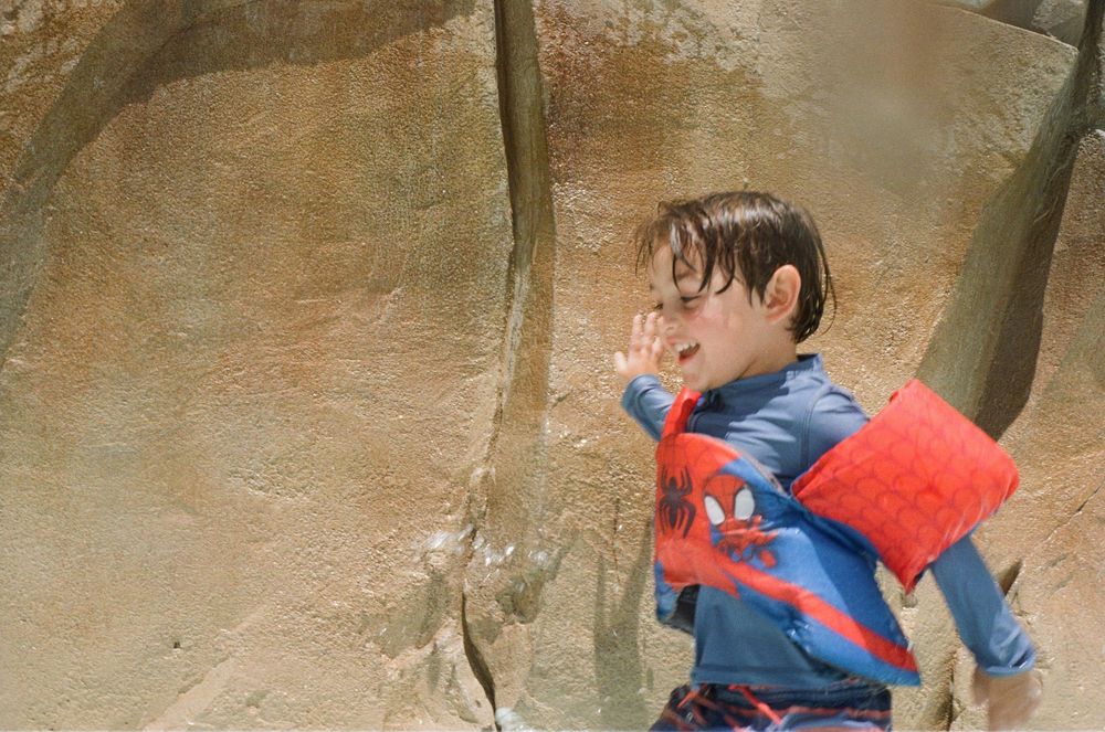 A young child in a Spider-Man costume plays outdoors against a tan rock wall background.