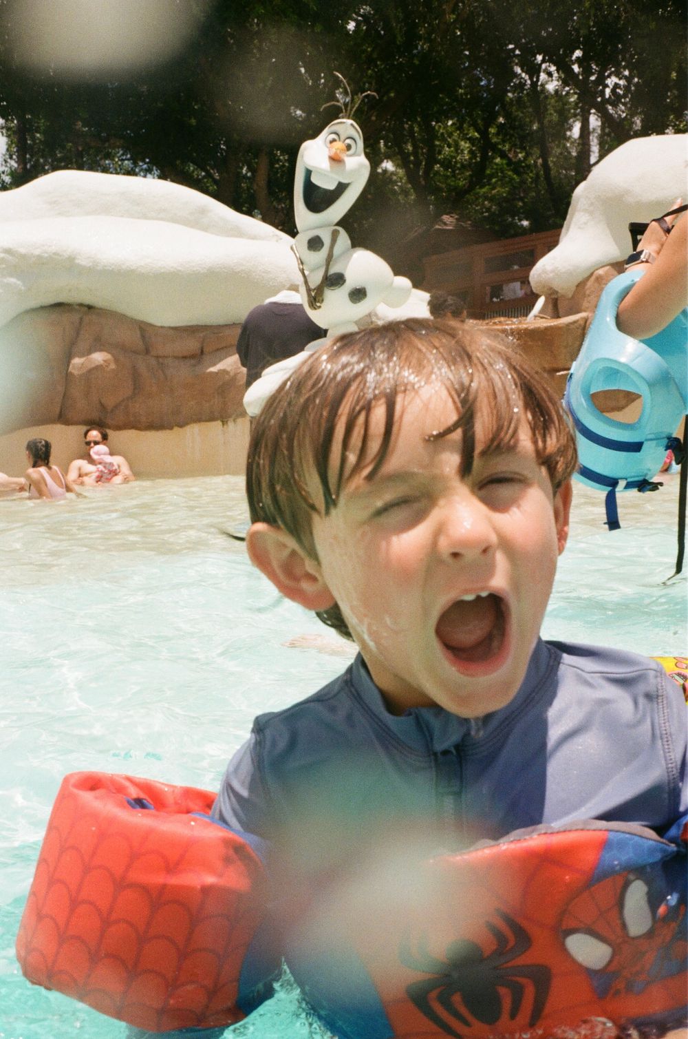 Child enjoying a splash in the pool while wearing a Spiderman floatation device and swim gear.