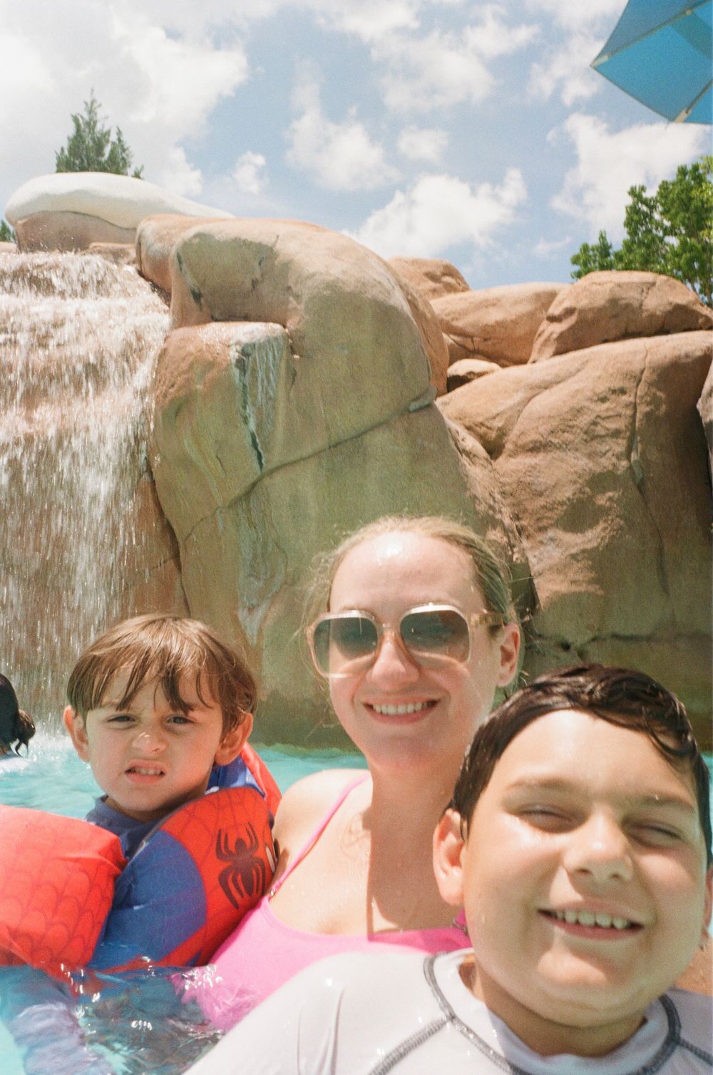 People enjoying a pool or water feature with rock formations and a waterfall at a water park on a sunny day.