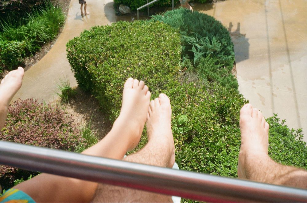 Two pairs of bare feet dangling over railing with green garden landscaping visible below in background.