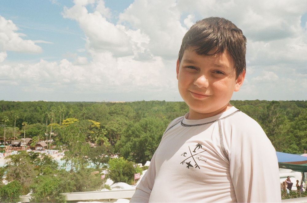 A young child in a white t-shirt stands on an elevated viewpoint overlooking a scenic outdoor landscape.