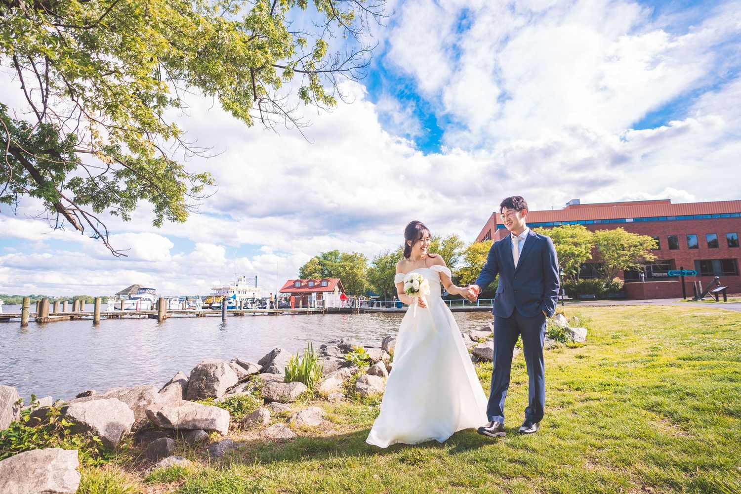 A couple shares their wedding moment on the grassy waterfront by a marina with boats and cloudy blue skies overhead.