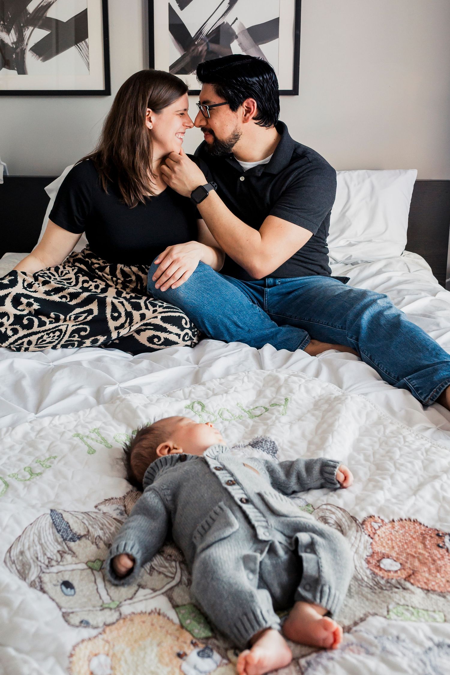 Young couple sitting on bed sharing an intimate moment while their baby lies sleeping in gray onesie nearby.