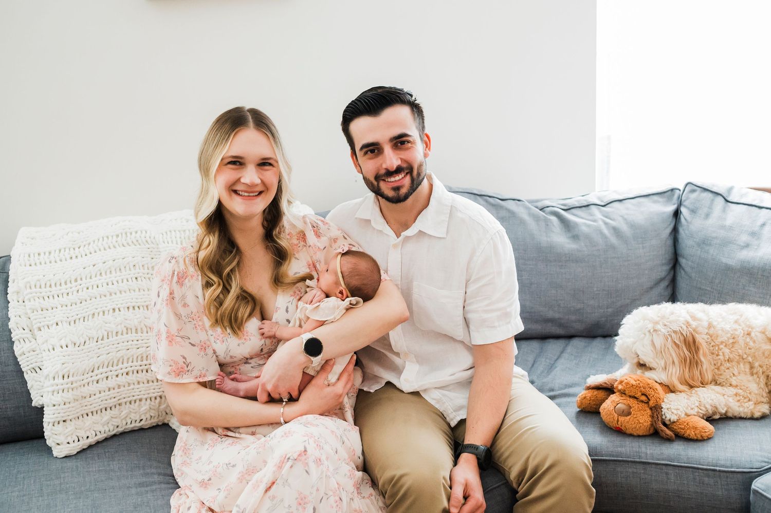 A loving family sits together on a gray couch with their sleeping dog nearby.
