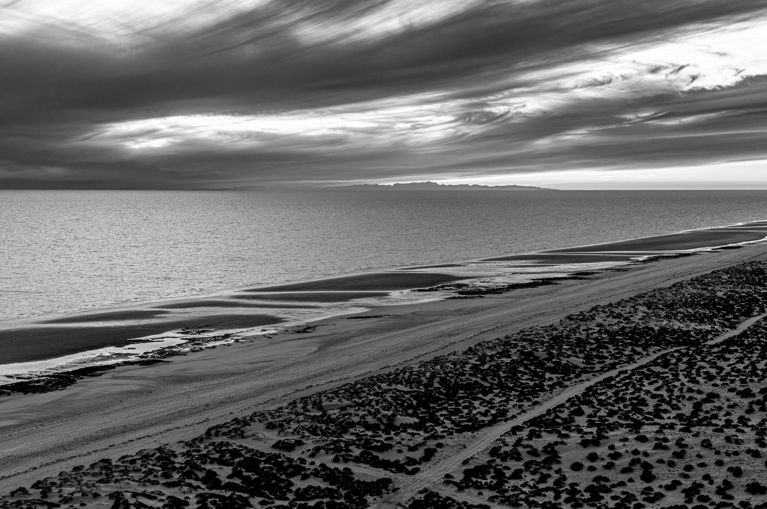 Dramatic black and white coastal landscape with sweeping clouds over shoreline.