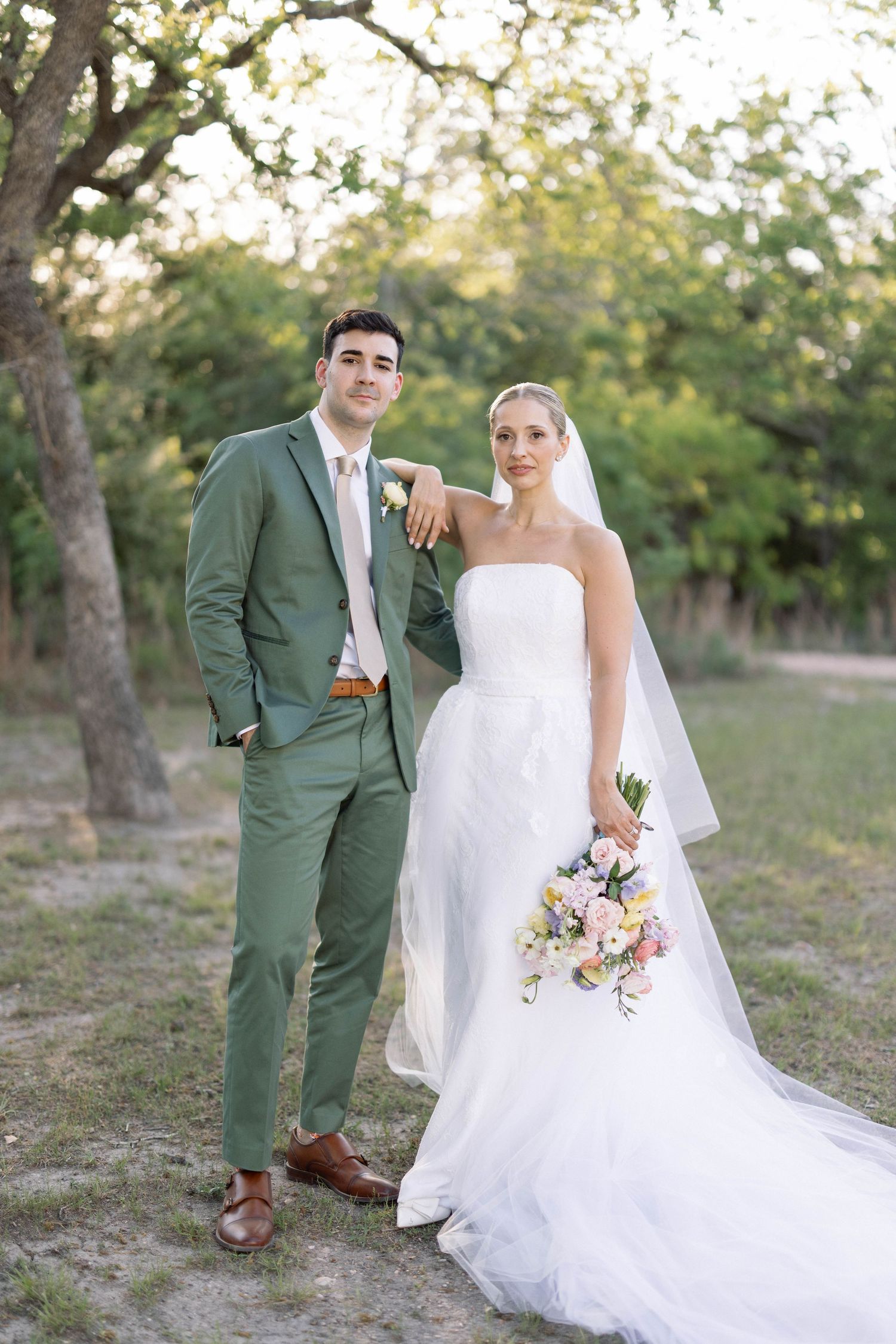 Wedding couple stands together in outdoor garden setting with bride wearing white gown and groom in sage green suit.