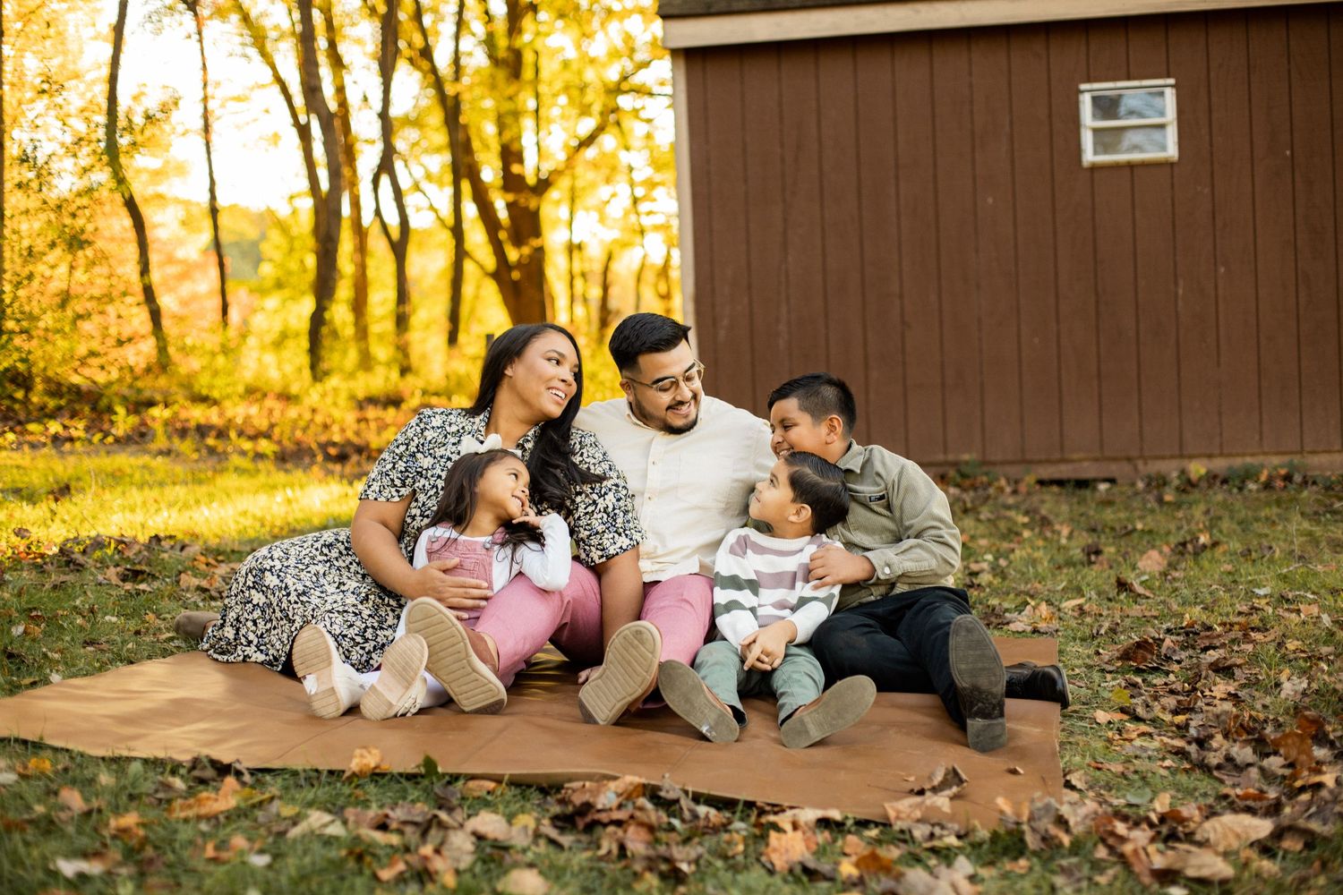 Family lounges together on blanket by brown building during fall photo session.