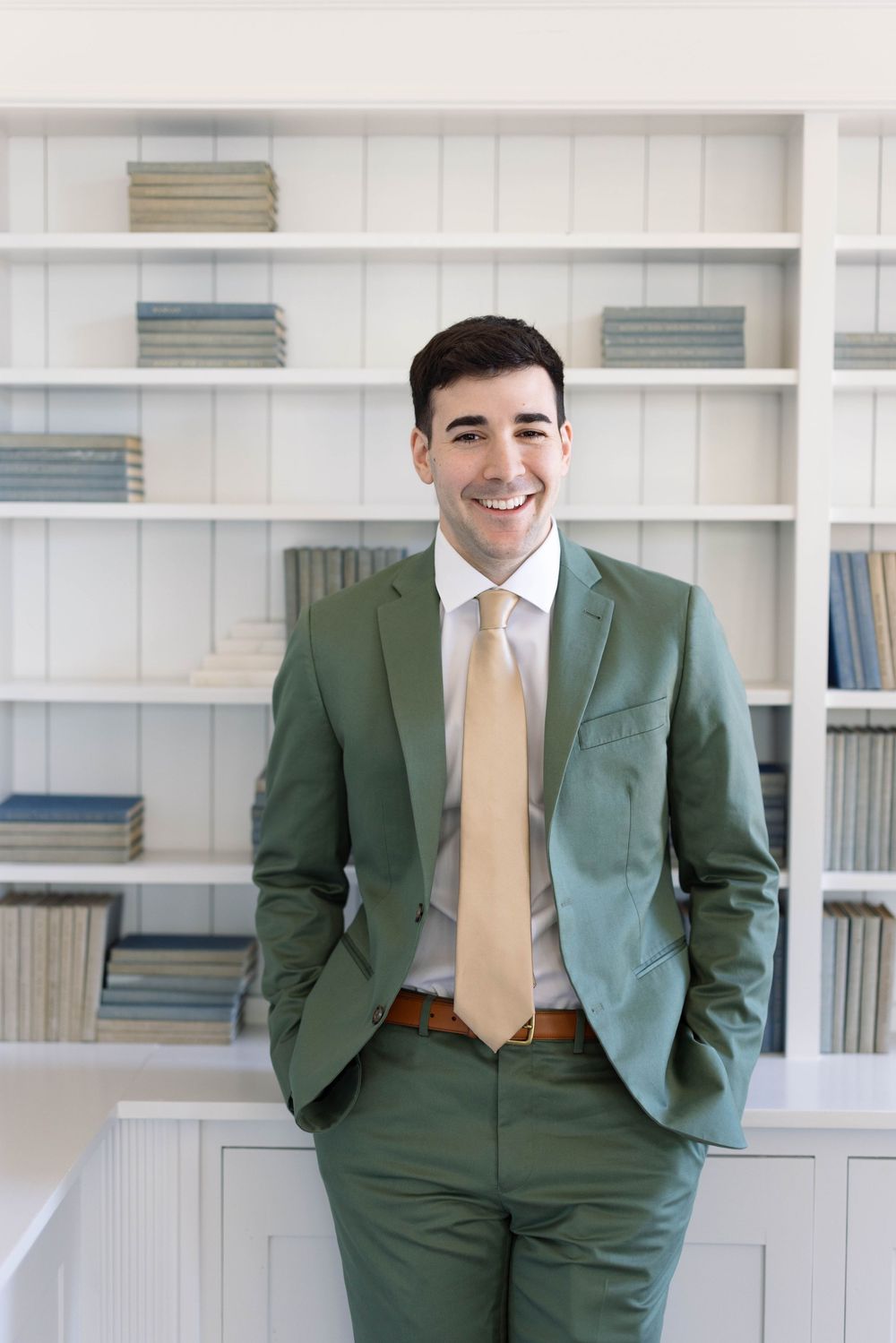 Professional in a stylish green suit and gold tie stands confidently against white built-in shelving with books.
